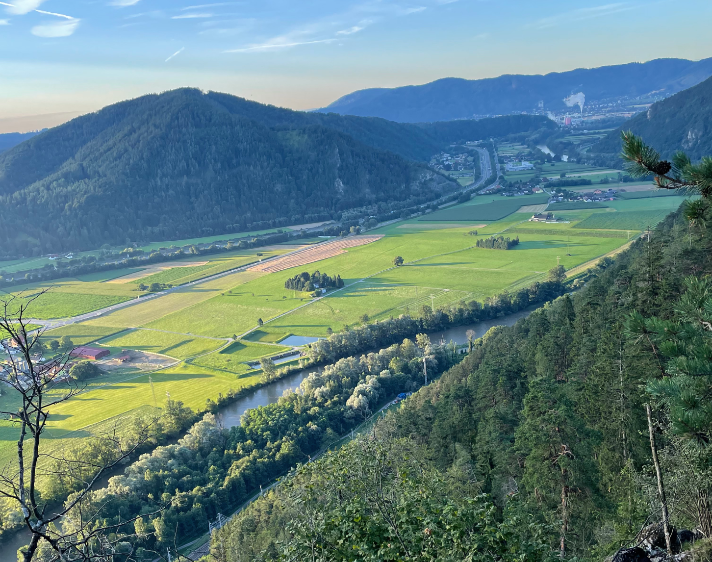 Panoramablick auf ein weites Tal. Ein Fluss schlängelt sich neben sonnenbeschienene grüne Felder, gesäumt von Straßen und Bahngleisen. Bewaldete Berge umrahmen die Szene. In der Ferne sind Siedlungen und Industriegebäude sichtbar.