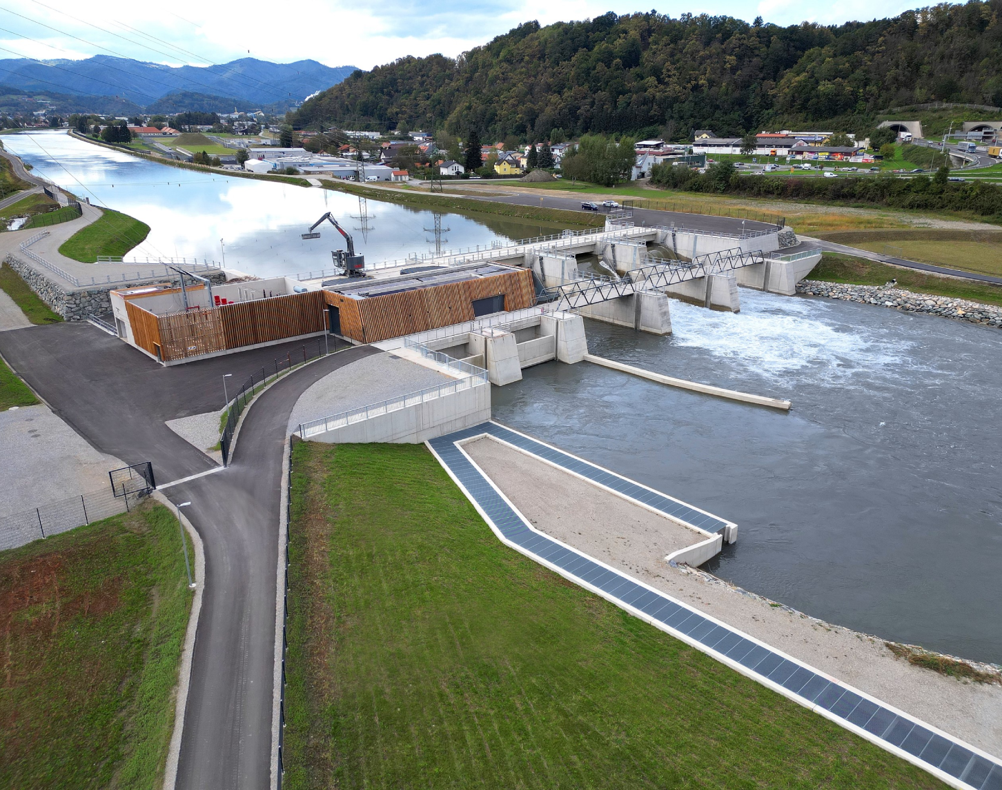 Ein modernes Wasserkraftwerk an einem Fluss in bergiger Landschaft, aus der Vogelperspektive. Vor dem Wehr liegt ruhiges Stauwasser, dahinter fließt das Wasser schäumend ab. Teile des Kraftwerkes sind noch nicht fertig und werden noch gebaut.