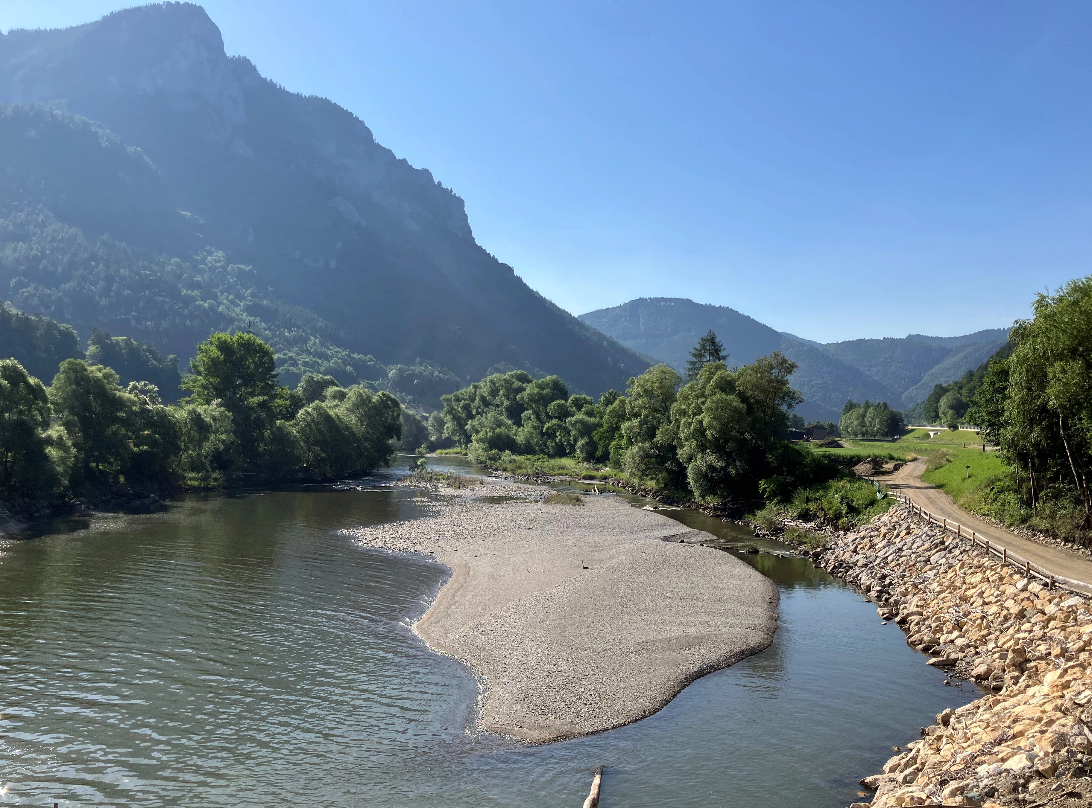 Ein breiter Fluss mit einer großen Kiesbank im Zentrum. Dicht bewaldete Ufer und Berge umgeben das Gewässer. Eine Straße läuft entland einem Teil des rechten Ufers. Am Horizont sind Häuser und ein Strommast erkennbar.